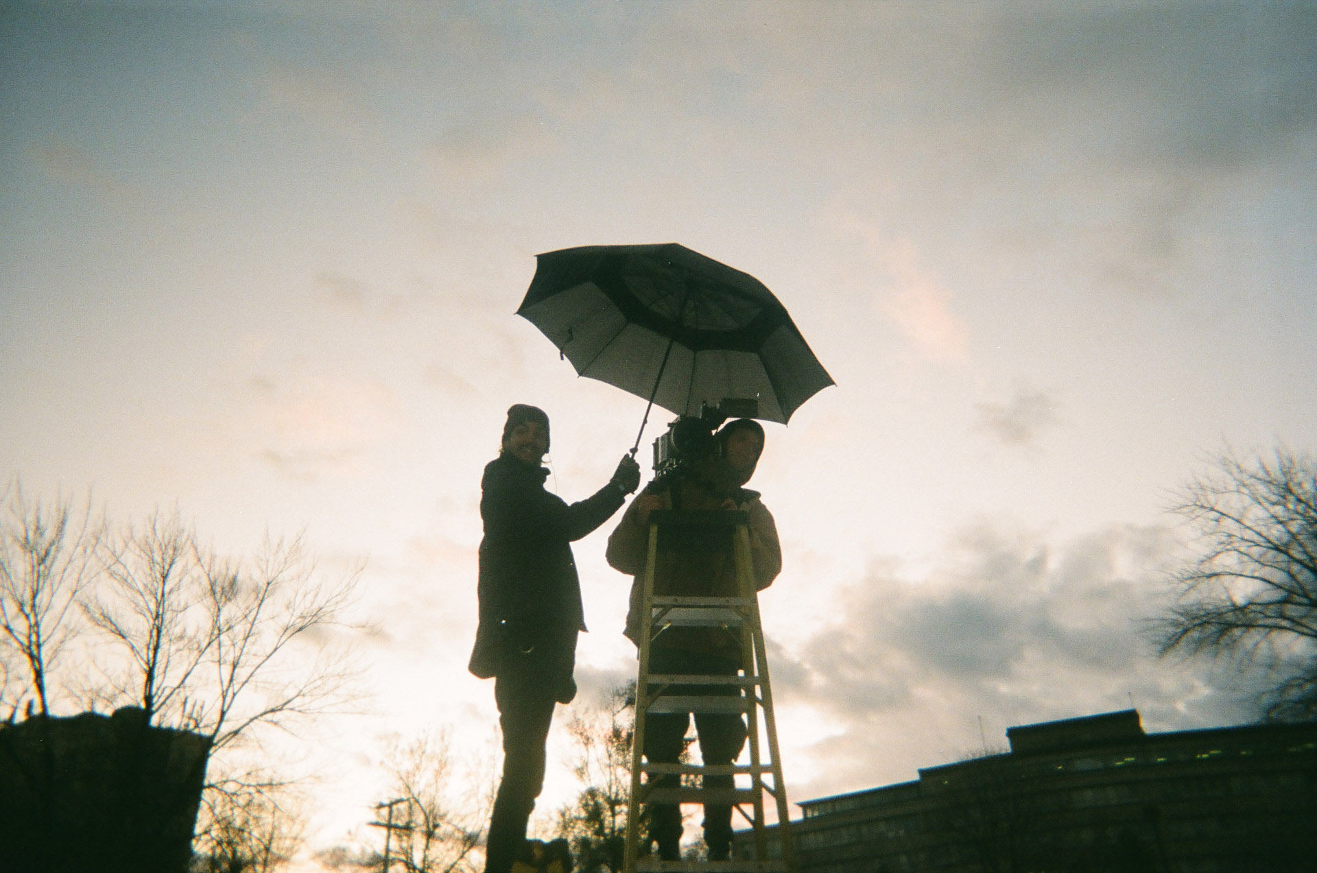 Vincent Ruel-Cote from Les Gamins up on high ladder with camera and assistant holding umbrella over him behind the scenes filming TV series La Loi C'est La Loi at night with TV5 Unis