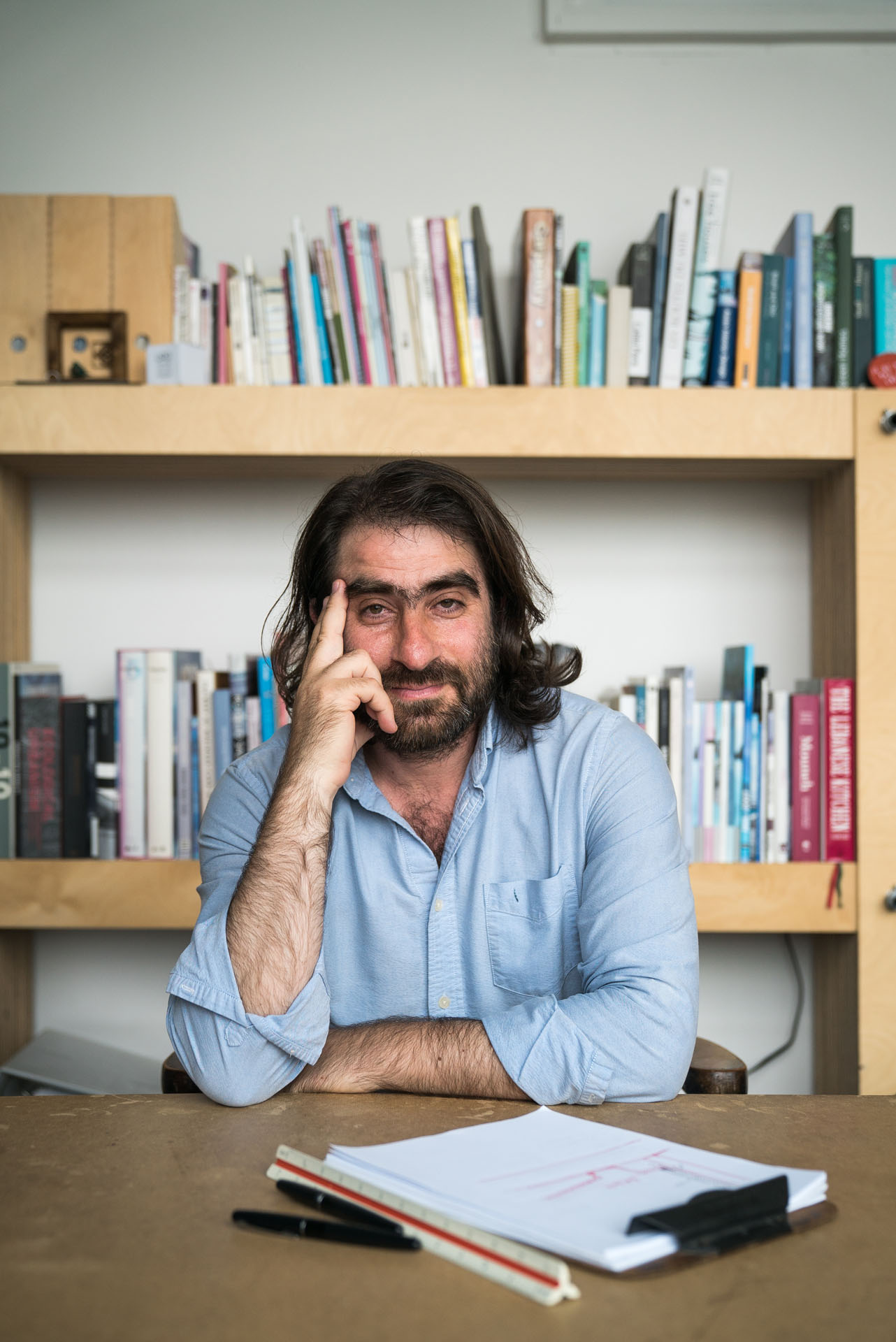 male writer long hair beard and unibrow sitting at desk in front of library looking at camera wearing light blue shirt by Bruno Florin for mile ex mille vie