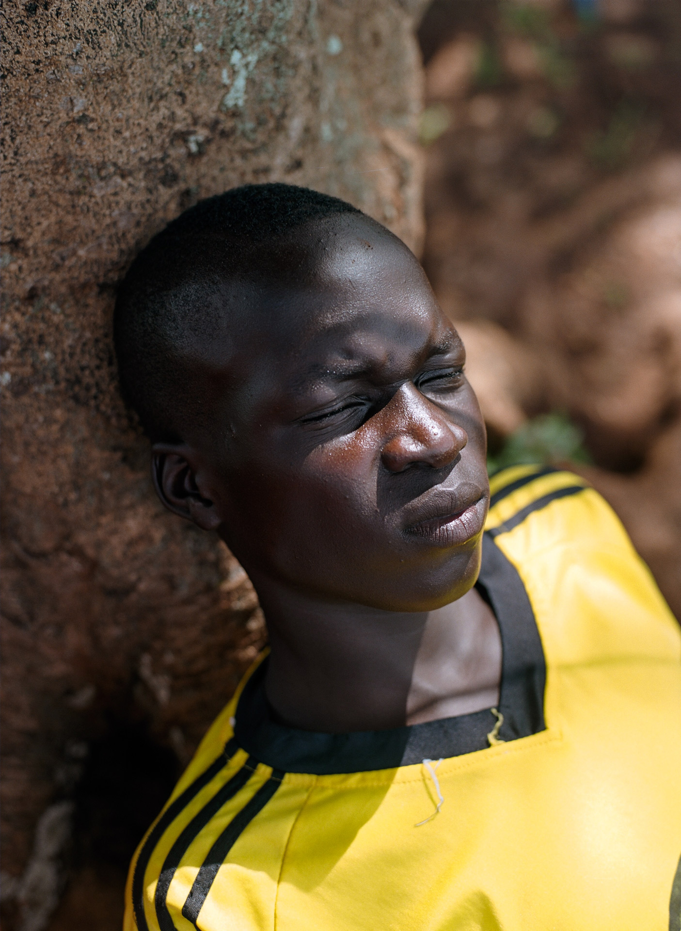young black boy resting in the shade frowning with eyes closed against a tree by Alexi Hobbs in Uganda for Football for good with Sportsnet