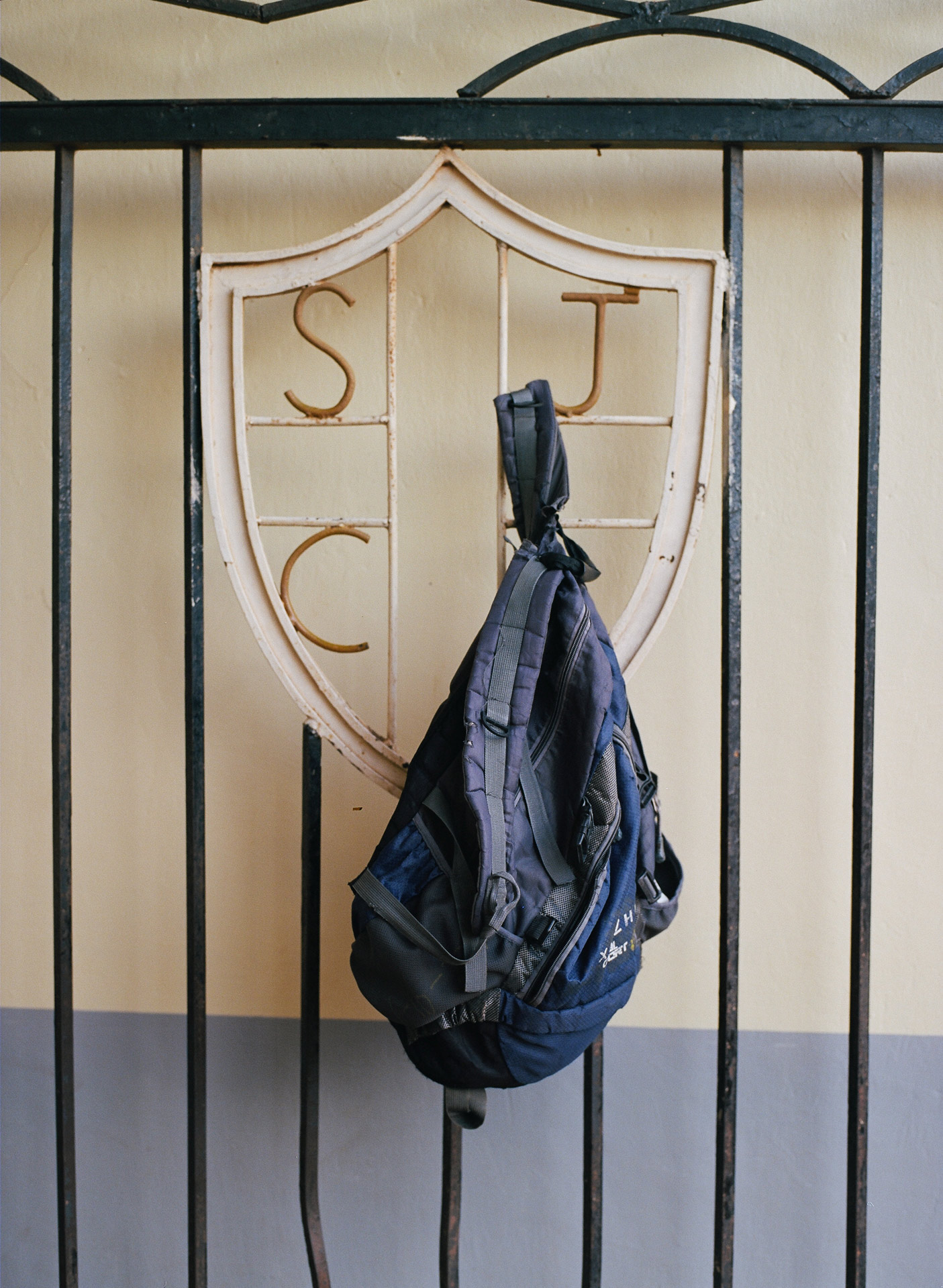 beaten up dark blue backpack hanging on metal fence with monogram by Alexi Hobbs in Uganda for Football for Good with Sportsnet
