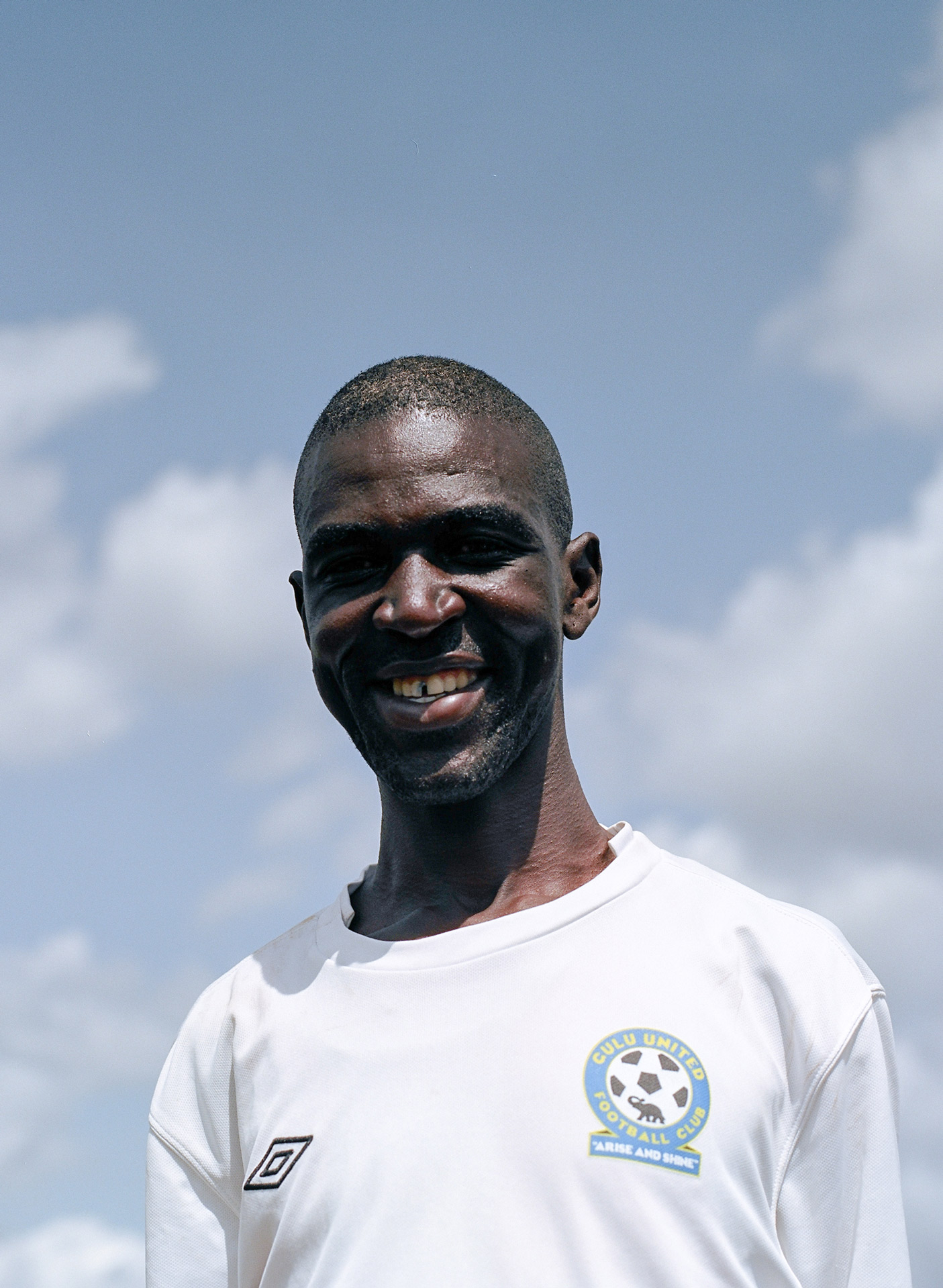 portrait of black man wearing white shirt smiling at camera with blue sky in the background by Alexi Hobbs in Uganda for Football for Good with Sportsnet