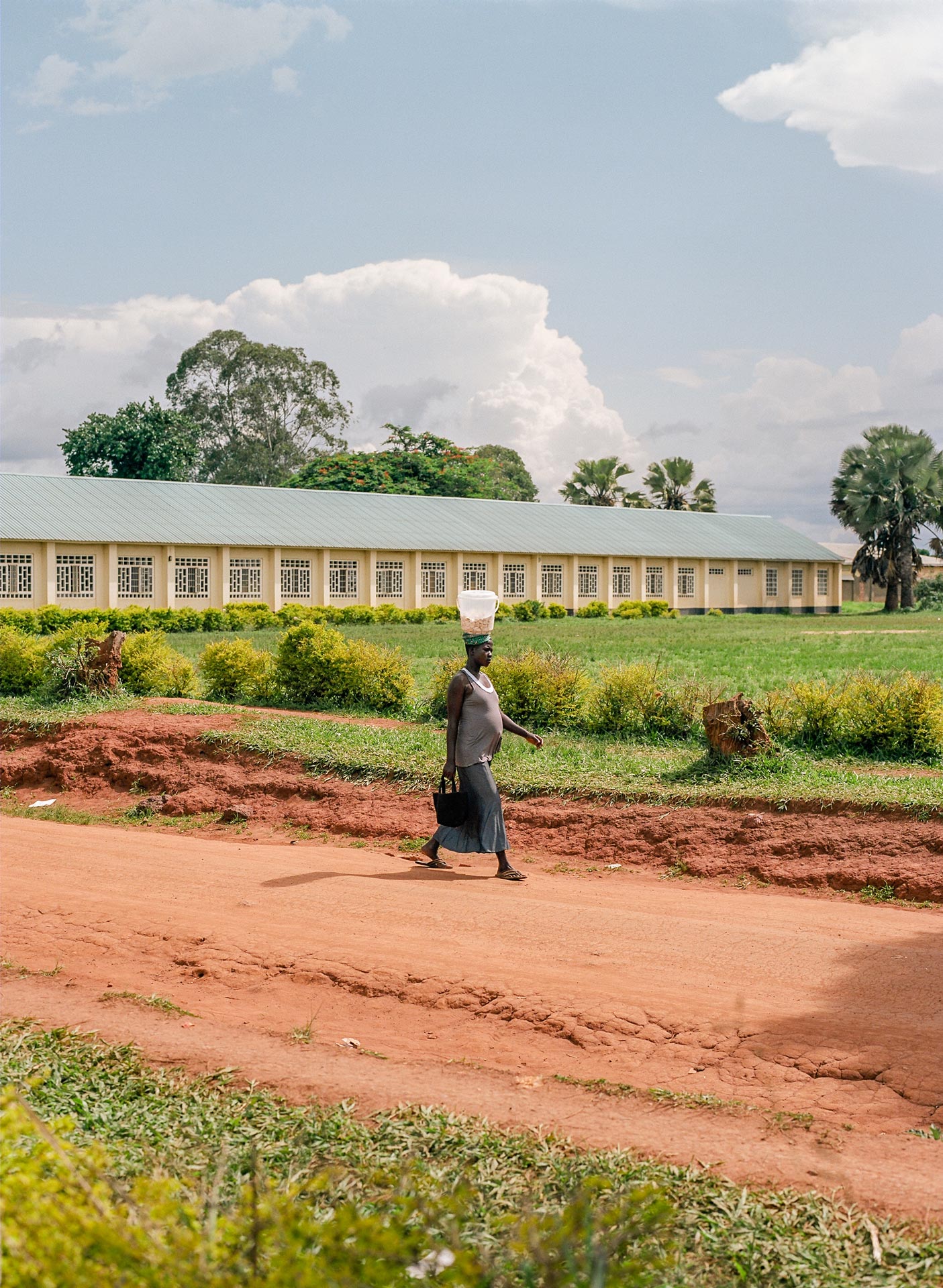 pregnant black woman walking on dirt road in the sun with bucket of rocks on her head by Alexi Hobbs in Uganda for Football for Good with Sportsnet