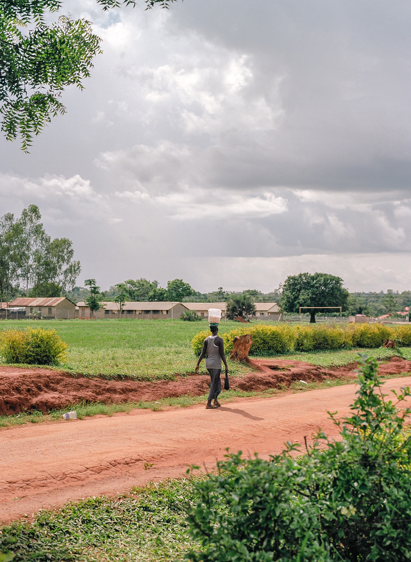 back of pregnant black woman walking on dirt road in the sun with bucket of rocks on her head by Alexi Hobbs in Uganda for Football for Good with Sportsnet