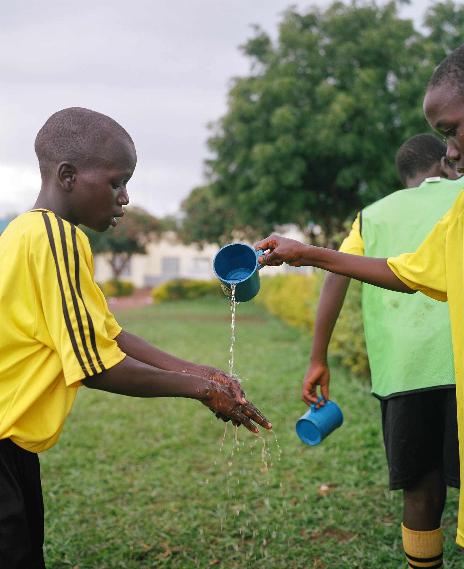 young black boy slowly pouring a blue cup of water for another boy to wash his hands by Alexi Hobbs in Uganda for Football for Good with Sportsnet