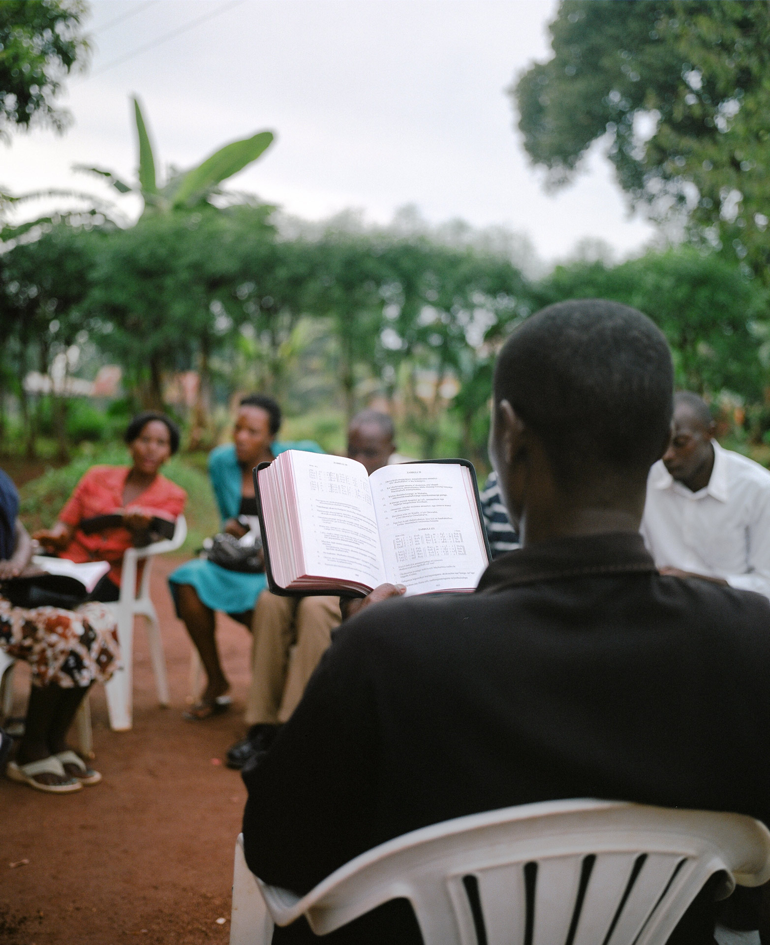 black man sitting on white plastic chair reading the bible with other people by Alexi Hobbs in Uganda for Football for Good with Sportsnet