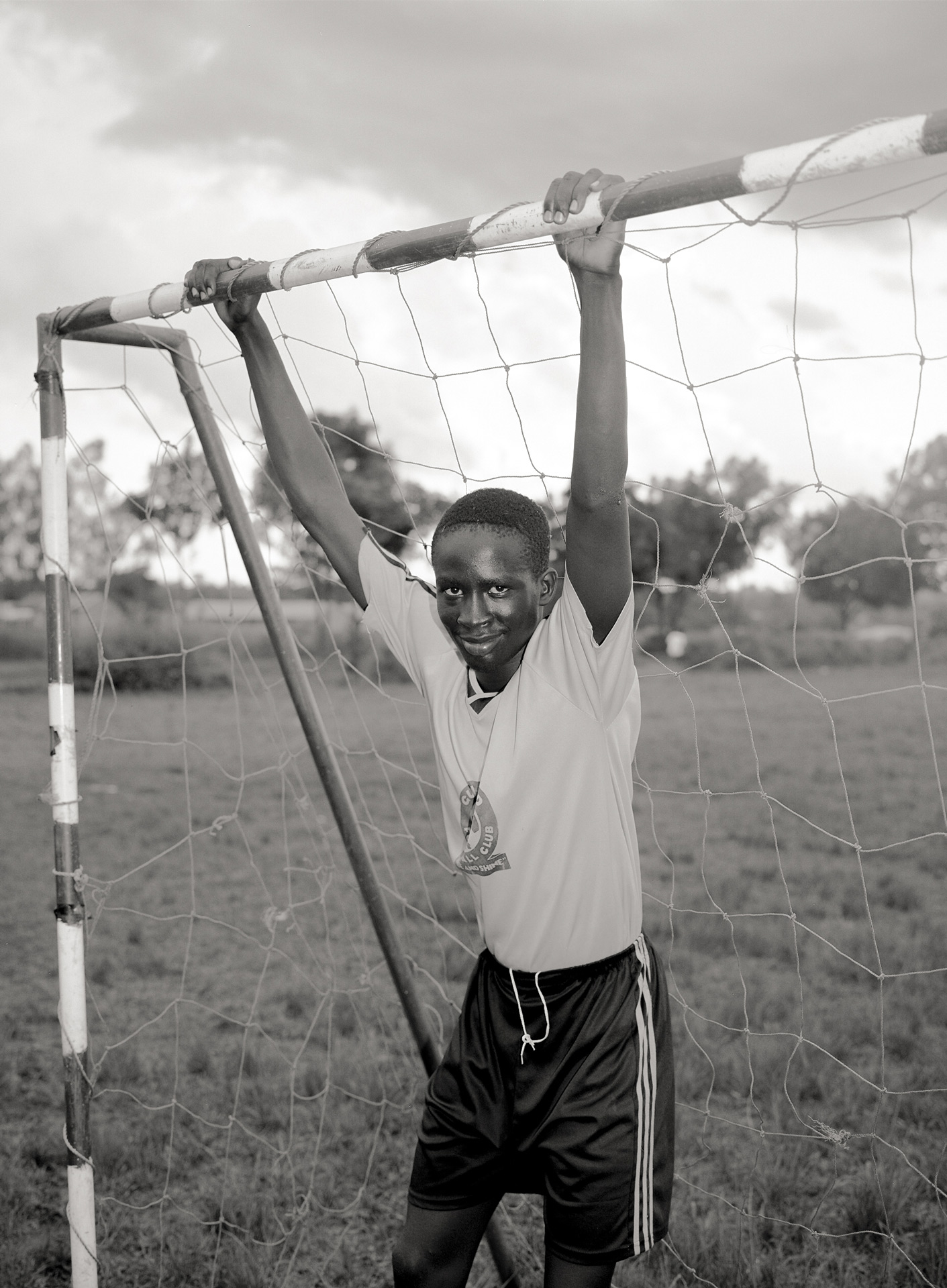 black and white picture of young goalie suspended by the metal frame looking at camera wearing yellow soccer jersey and black sports short by Alexi Hobbs in Uganda for Football for Good with Sportsnet