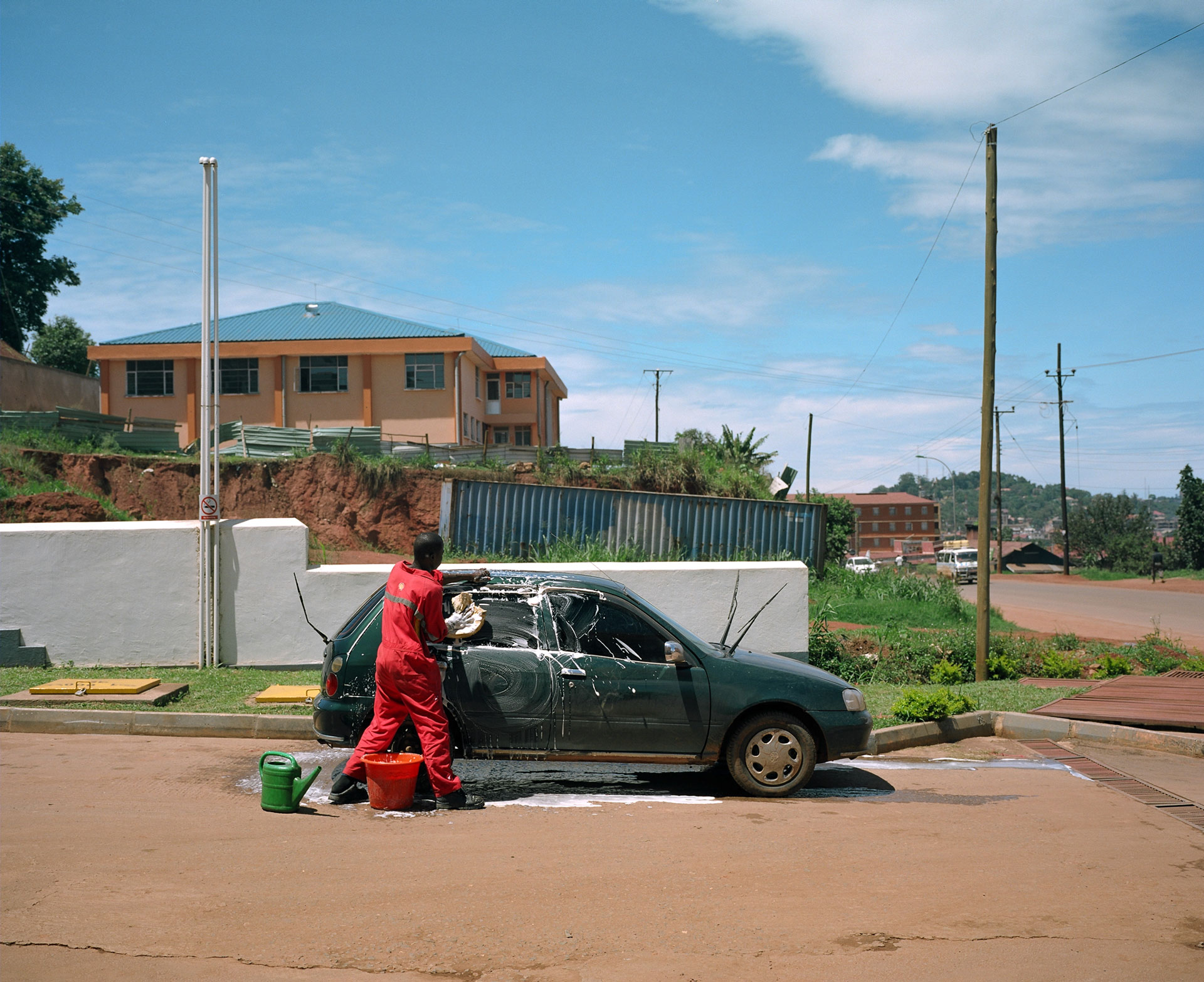 black man all dressed in red washing a dark green car by hand in the sun by Alexi Hobbs in Uganda for Football for Good with Sportsnet