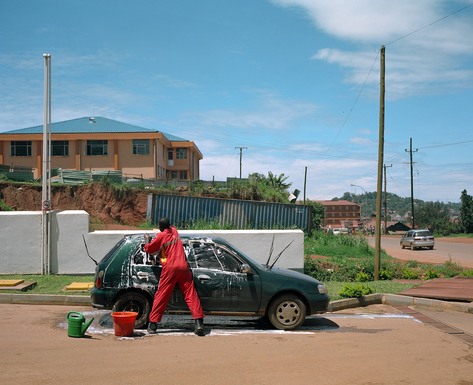 black man all dressed in red washing a dark green car by hand in the sun by Alexi Hobbs in Uganda for Football for Good with Sportsnet