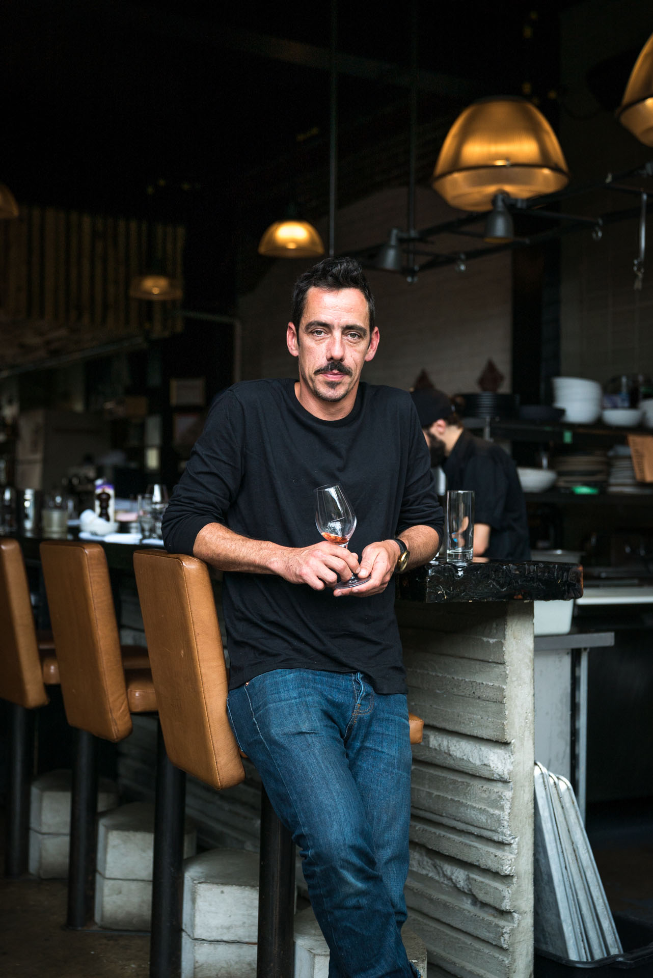 middle aged man standing against bar stool holding almost empty glass of wine by bruno florin for mile-ex mille vies