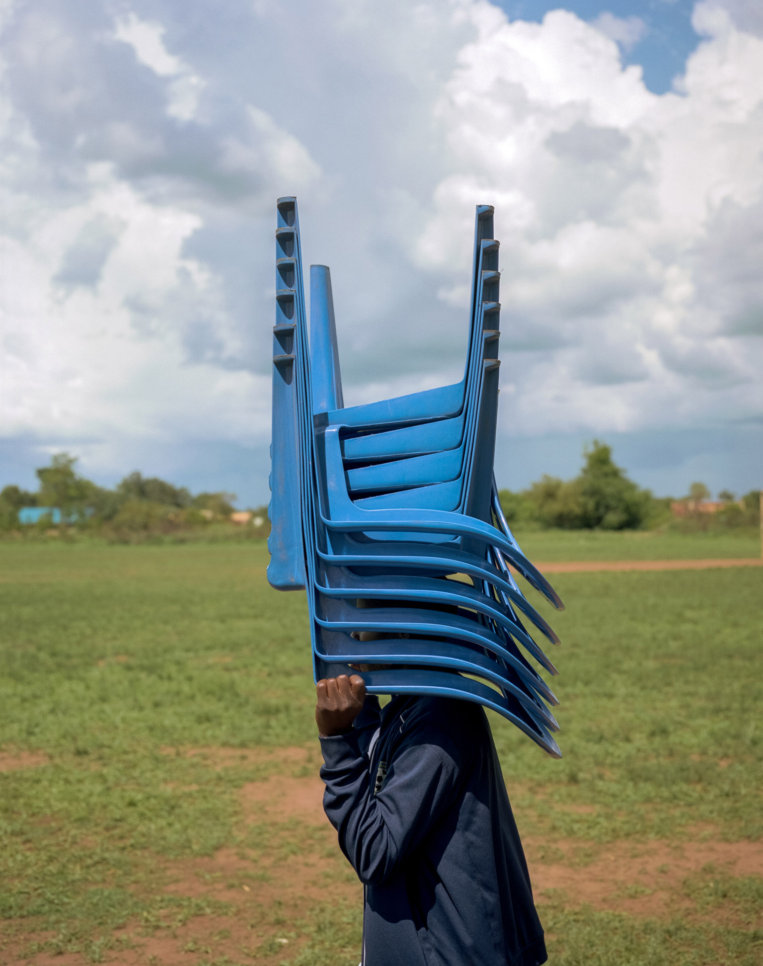young black boy transporting a pile of blue plastic chairs upside down on his head by Alexi Hobbs in Uganda for Football for good with Sportsnet