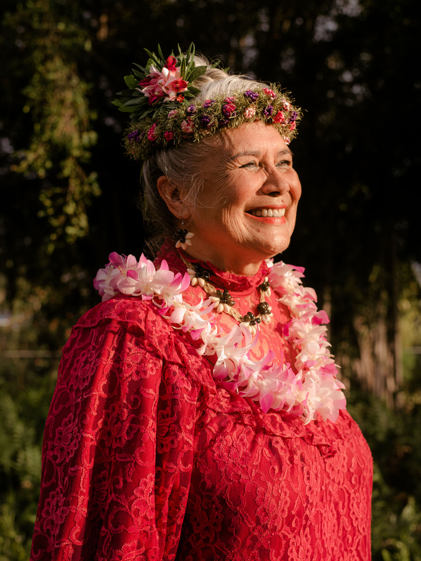 older lady wearing red lace traditional dress flower necklace flower crown smiling in the sun by Alexi Hobbs in Hawai'i for enRoute Magazine