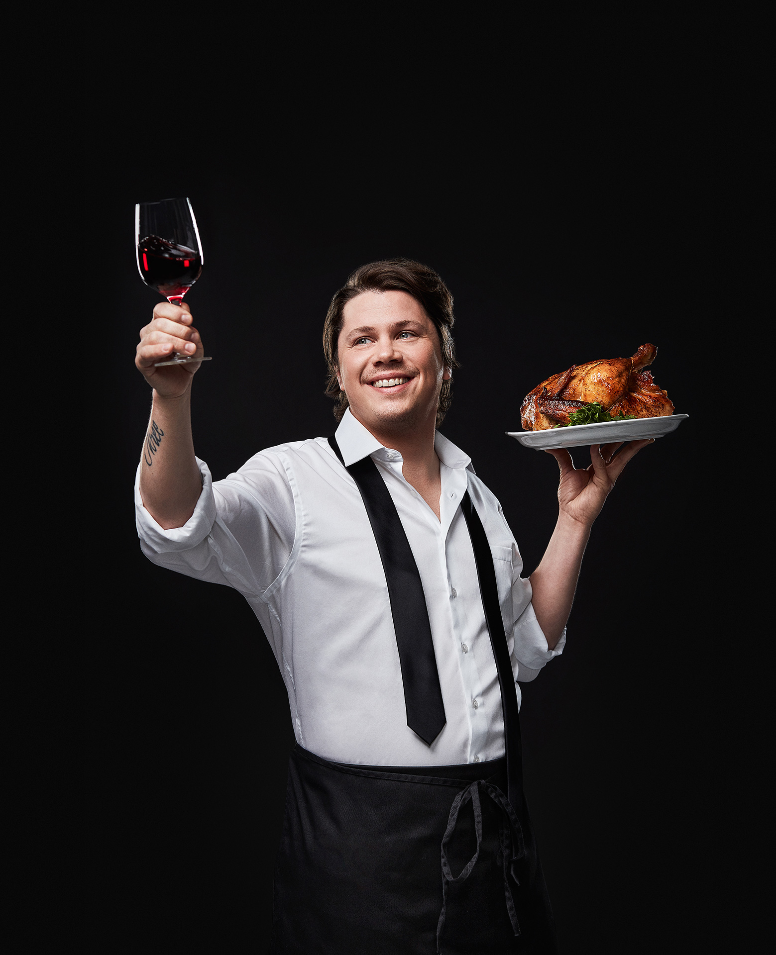 portrait of male chef Nicholas Giambattisto holding a glass of red wine in the air in one hand and a full roasted chicken on a plate in the other by Jocelyn Michel for the Cathcart