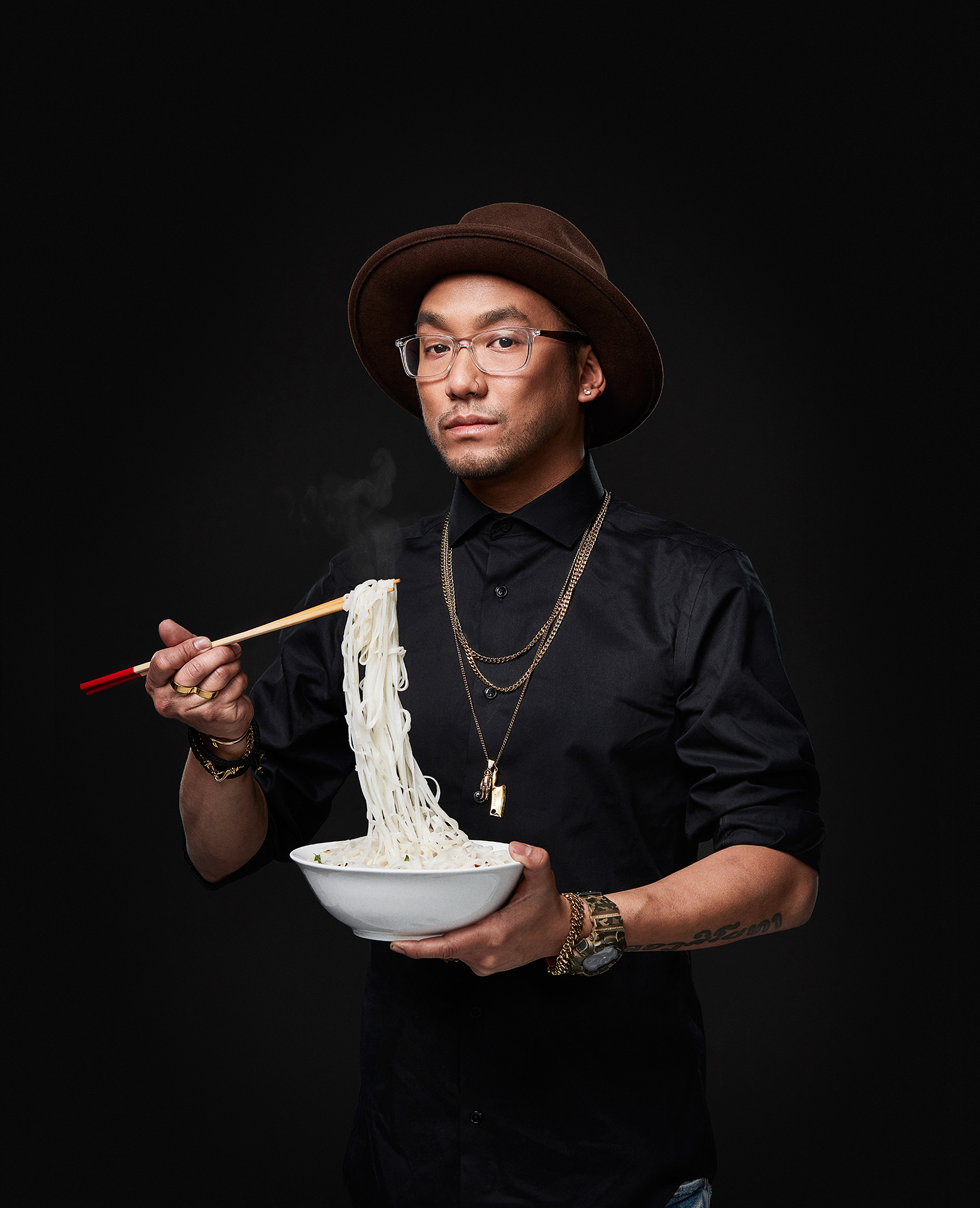 portrait of chef Ross Louangsignotha looking poised wearing jewelry holding chopsticks and a bowl moving rice noodles towards his mouth by Jocelyn Michel for the Cathcart