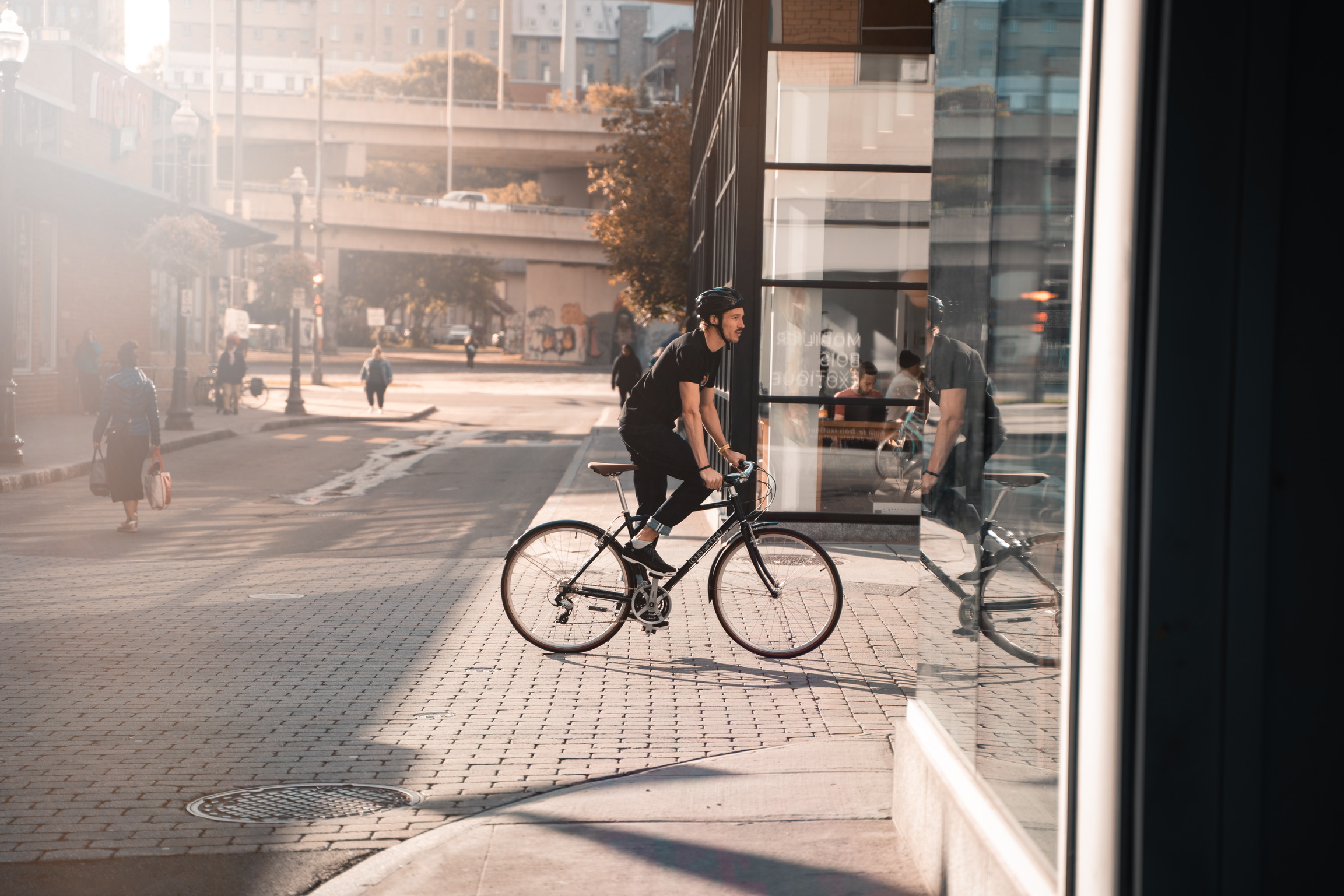 male model wearing black shirt with black jeans and black shoes biking in Montreal street on black bike in the sun as Spring Summer 2020 campaign of Louis Garneau with artistic direction and styling by Studio TB