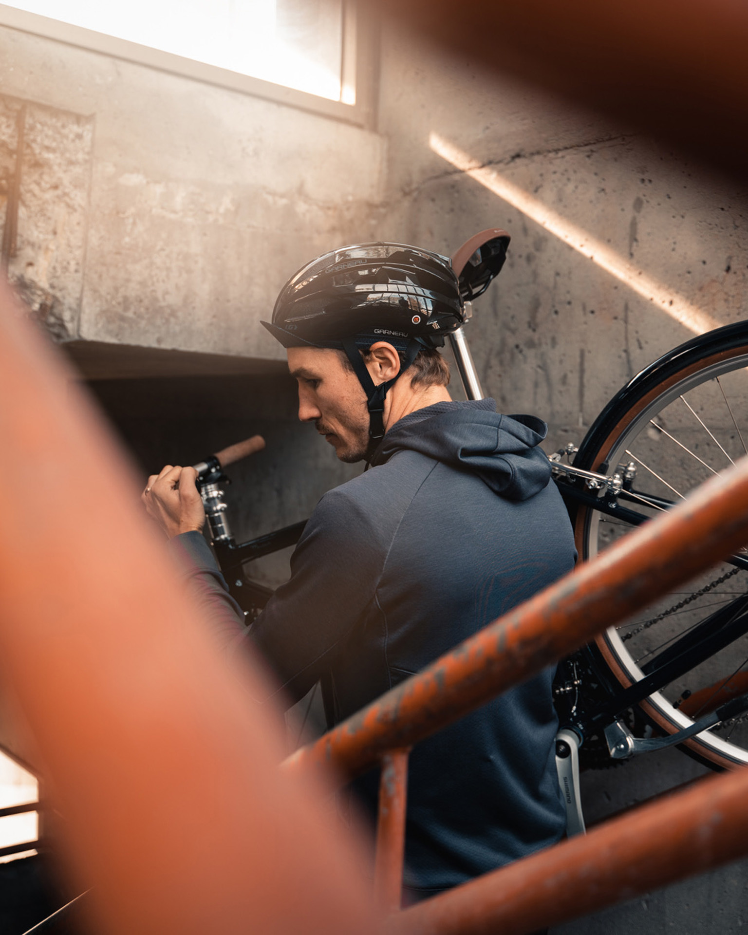 male model wearing bike helmet black shorts and dark blue jacket holding a bike over his shoulder while going down the stairs for Spring Summer 2020 campaign of Louis Garneau with artistic direction and styling by Studio TB