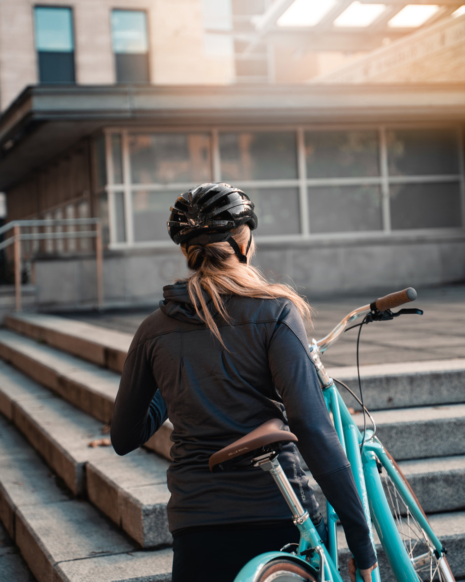 blonde female model wearing black bike helmet black shorts grey jacket and grey shoes hoisting up turquoise bike while walking up stairs for Spring Summer 2020 campaign of Louis Garneau with artistic direction and styling by Studio TB