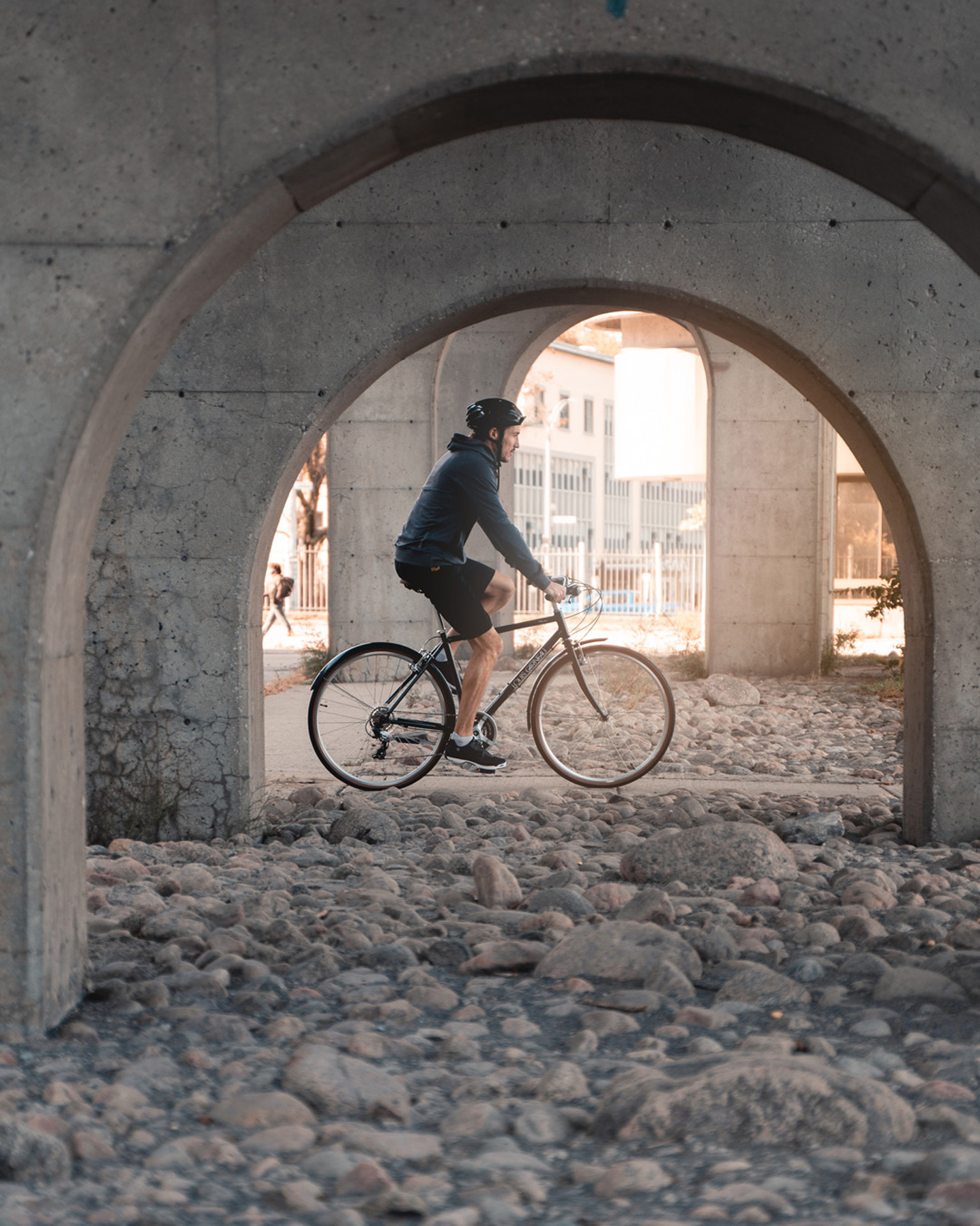 male model wearing bike helmet black shorts and dark blue jacket biking under a bridge on black bike as Spring Summer 2020 campaign of Louis Garneau with artistic direction and styling by Studio TB