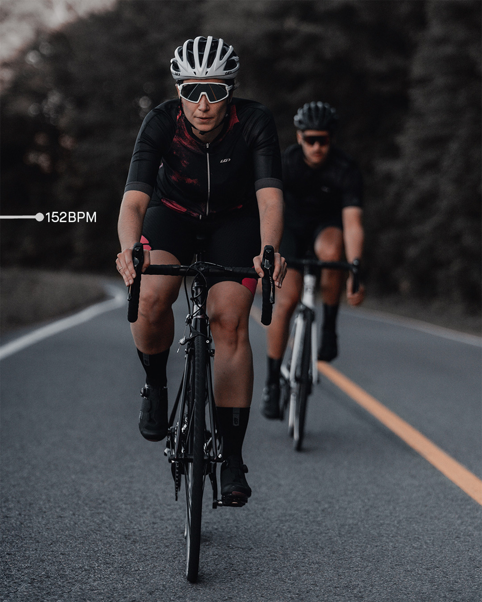 female model wearing black bike helmet and black and pink biking bodysuit with sunglasses biking on road on a black sports bike for Spring Summer 2020 campaign of Louis Garneau with artistic direction and styling by Studio TB