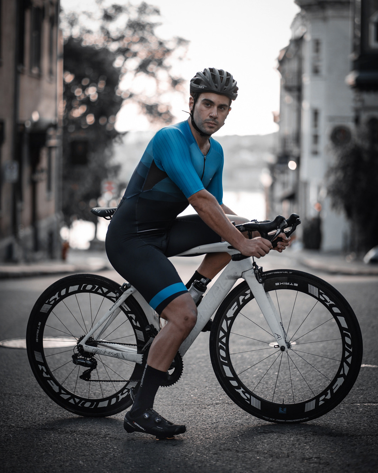 male model wearing black bike helmet and black and blue biking bodysuit sitting on a white sports bike for Spring Summer 2020 campaign of Louis Garneau with artistic direction and styling by Studio TB