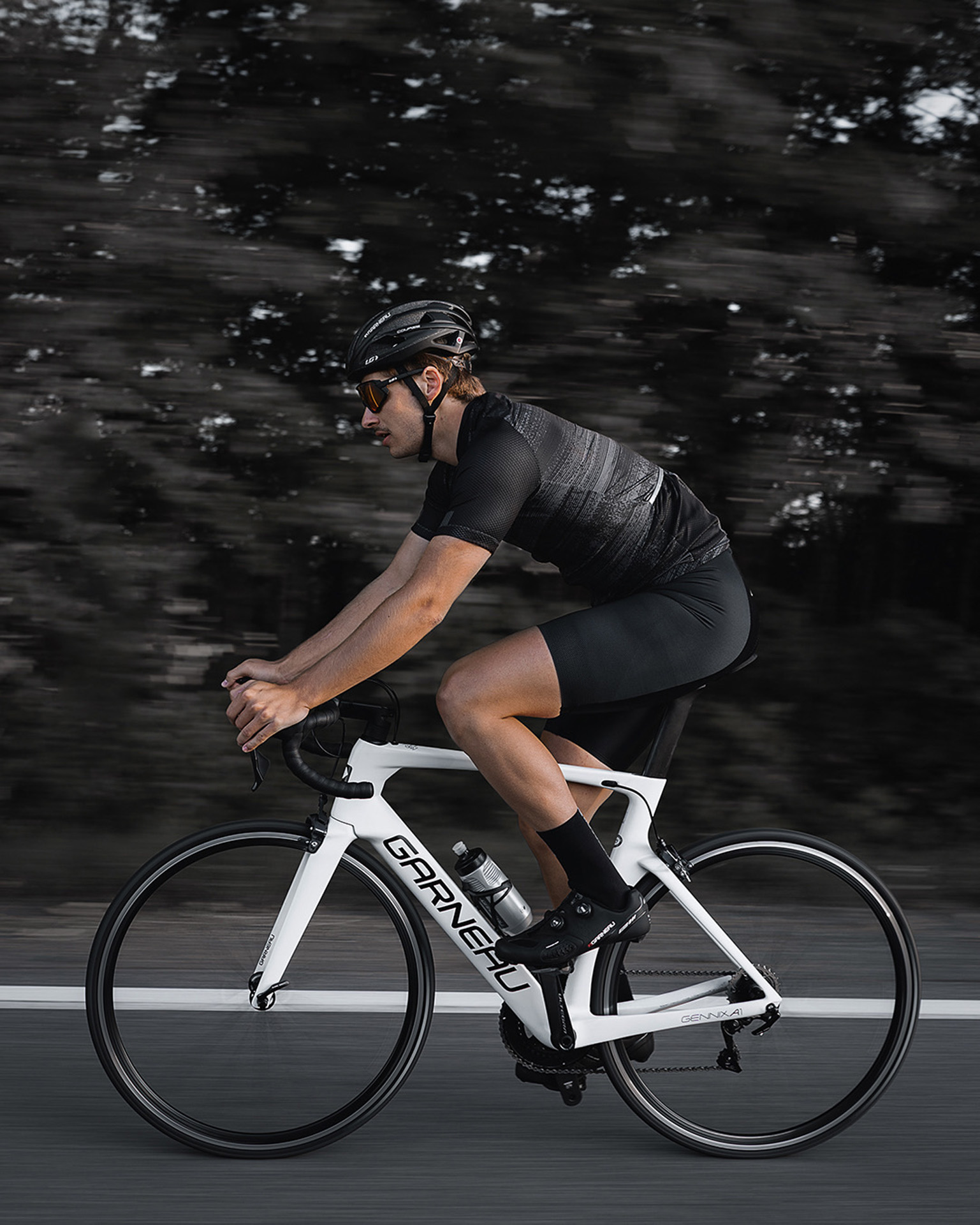 male model wearing black bike helmet and black biking bodysuit with sunglasses biking fast on road on a white sports bike for Spring Summer 2020 campaign of Louis Garneau with artistic direction and styling by Studio TB