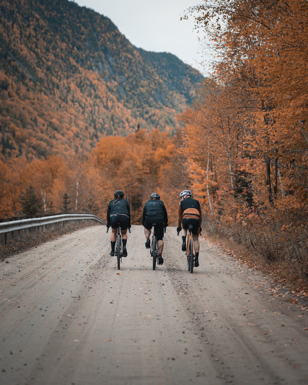 trio of long distance bikers biking on dirt road side by side with mountains covered in orange colored trees in the background for Spring Summer 2020 campaign of Louis Garneau with artistic direction and styling by Studio TB