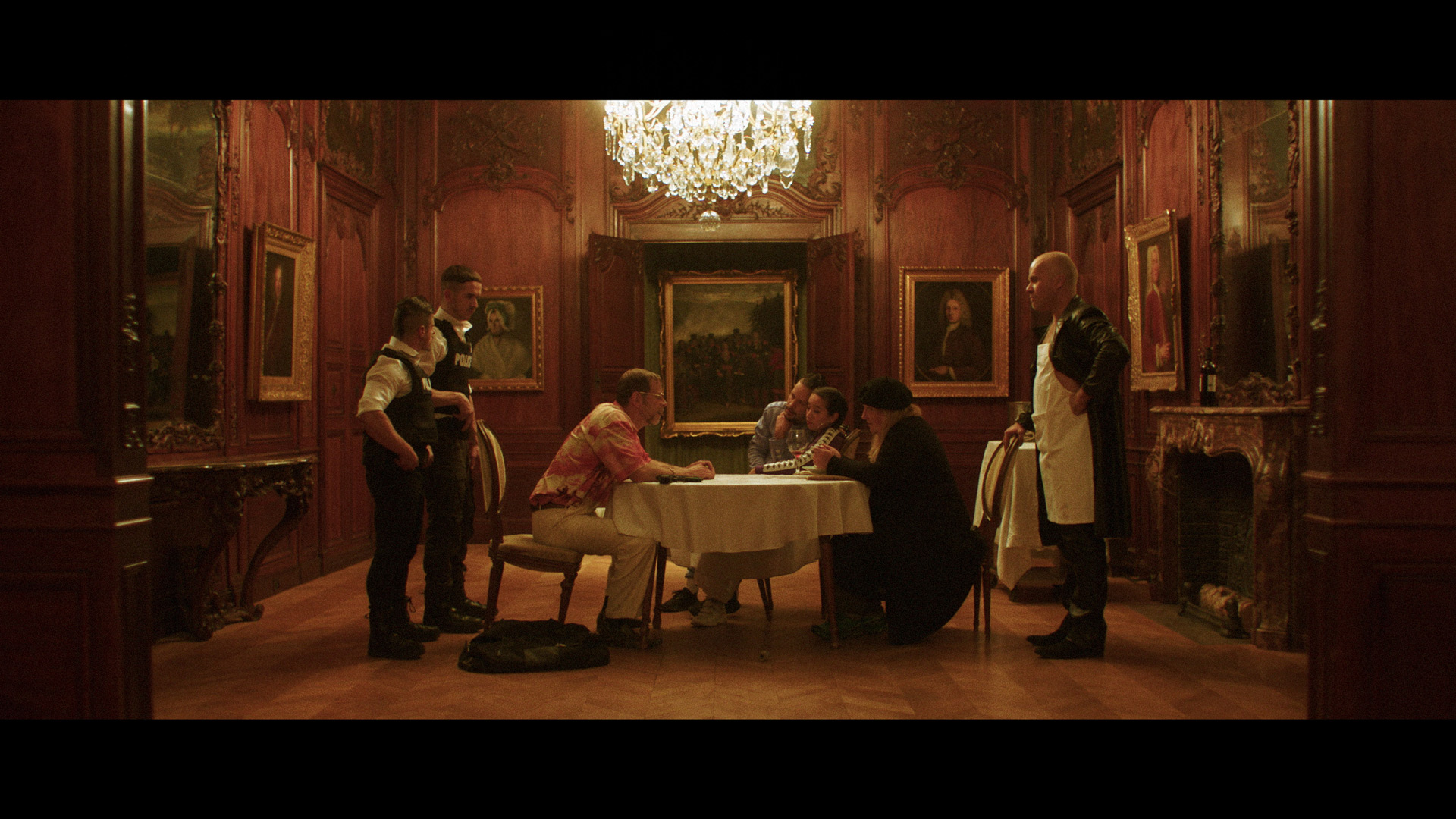 actors Hugolin Chevrette-Landesque and Emile Schneider posing as cops on each side of a man in tie-dye shirt holding a blue lollipop sitting at table in front of group of people for negotiations for TV series La Loi C'est La Loi filmed by Les Gamins for TV5 Unis