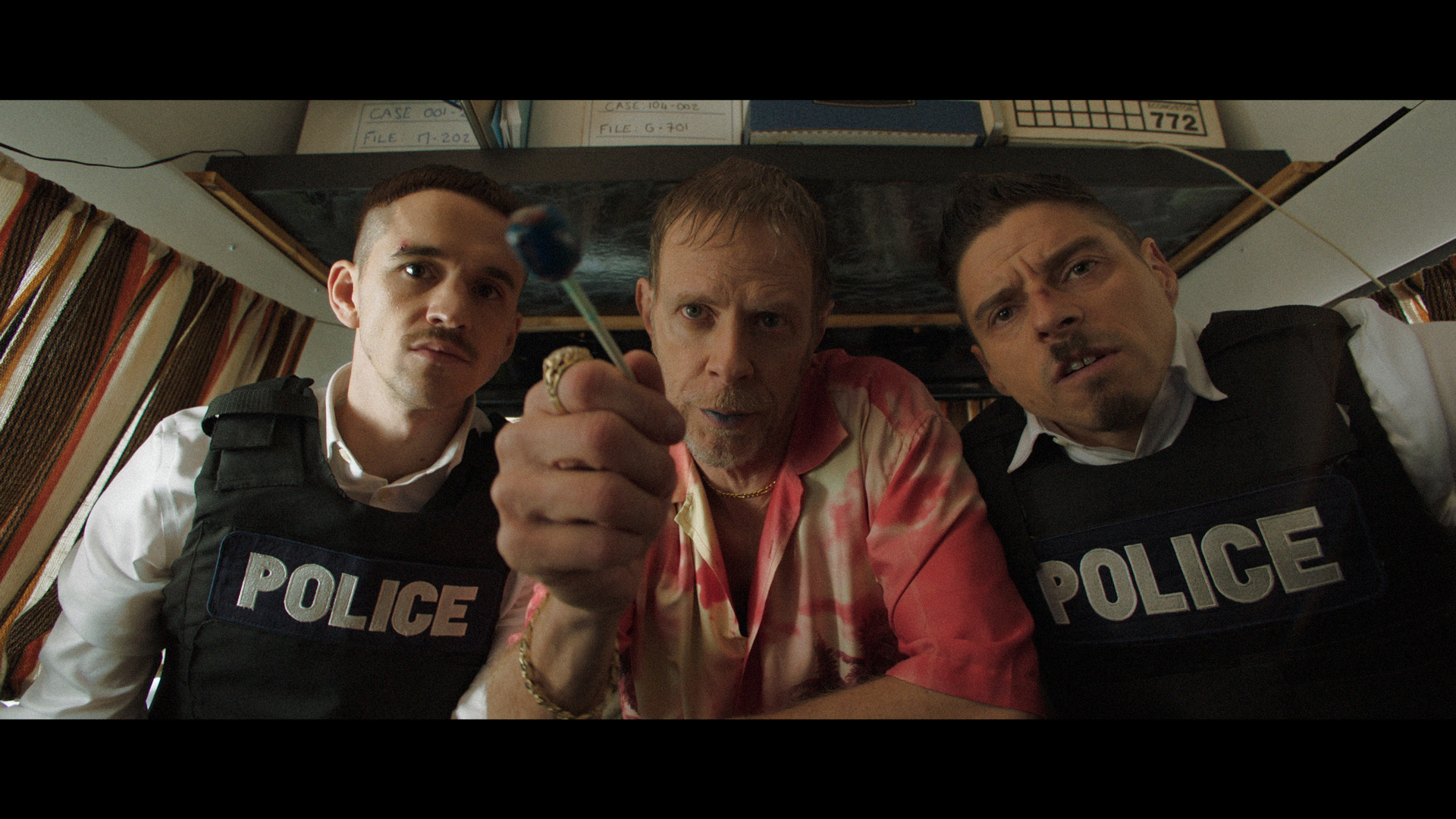 actors Hugolin Chevrette-Landesque and Emile Schneider posing as cops on each side of a man in tie-dye shirt holding a blue lollipop with blue lips looking at camera for TV series La Loi C'est La Loi filmed by Les Gamins for TV5 Unis