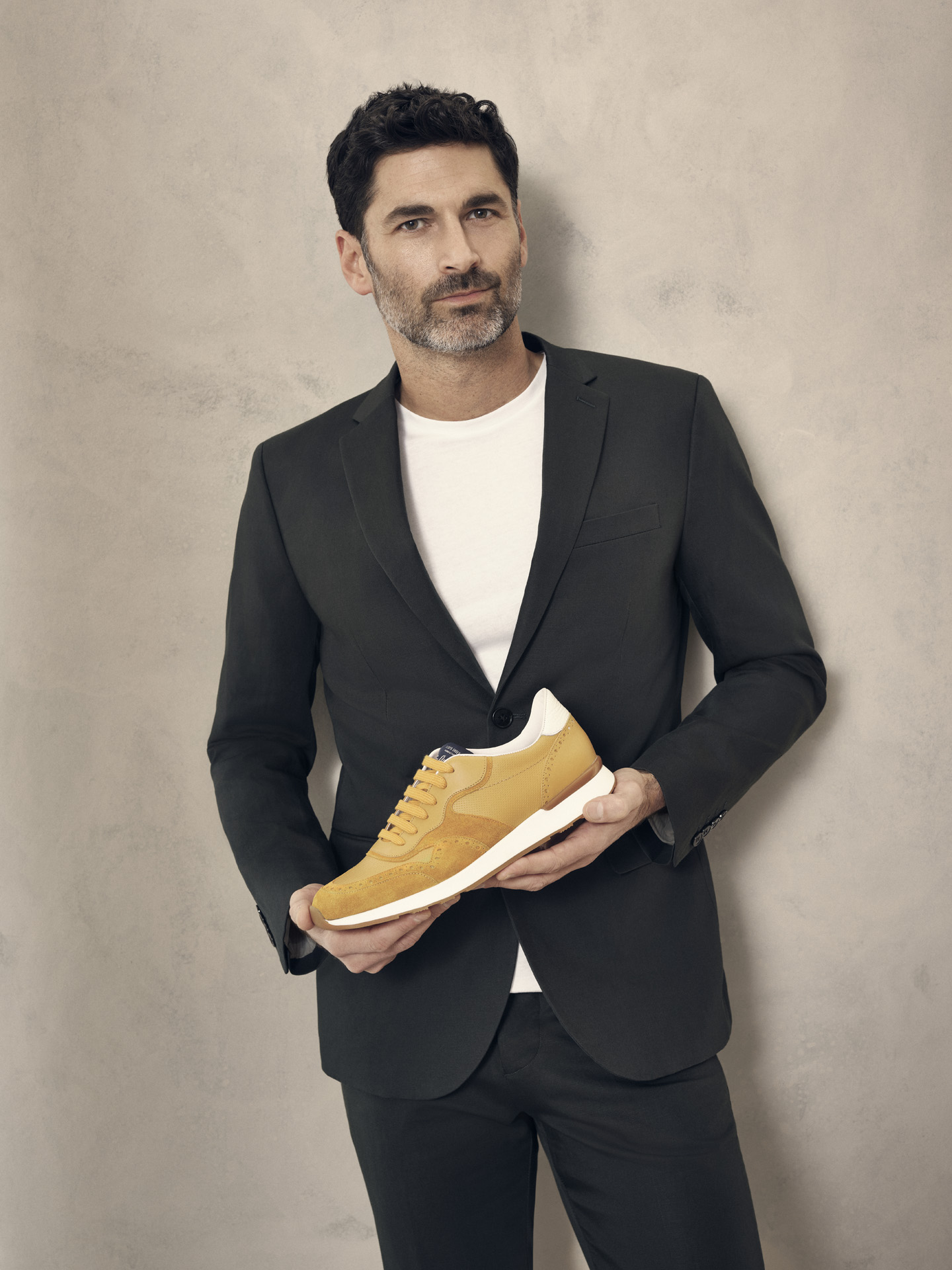 portrait of middle aged man wearing white t-shirt with black suit jacket and formal black pants holding a yellow sneaker for Jean-Paul Fortin summer-spring collection photographed by Maxyme G Delisle with artistic direction, conception and set design by Studio TB