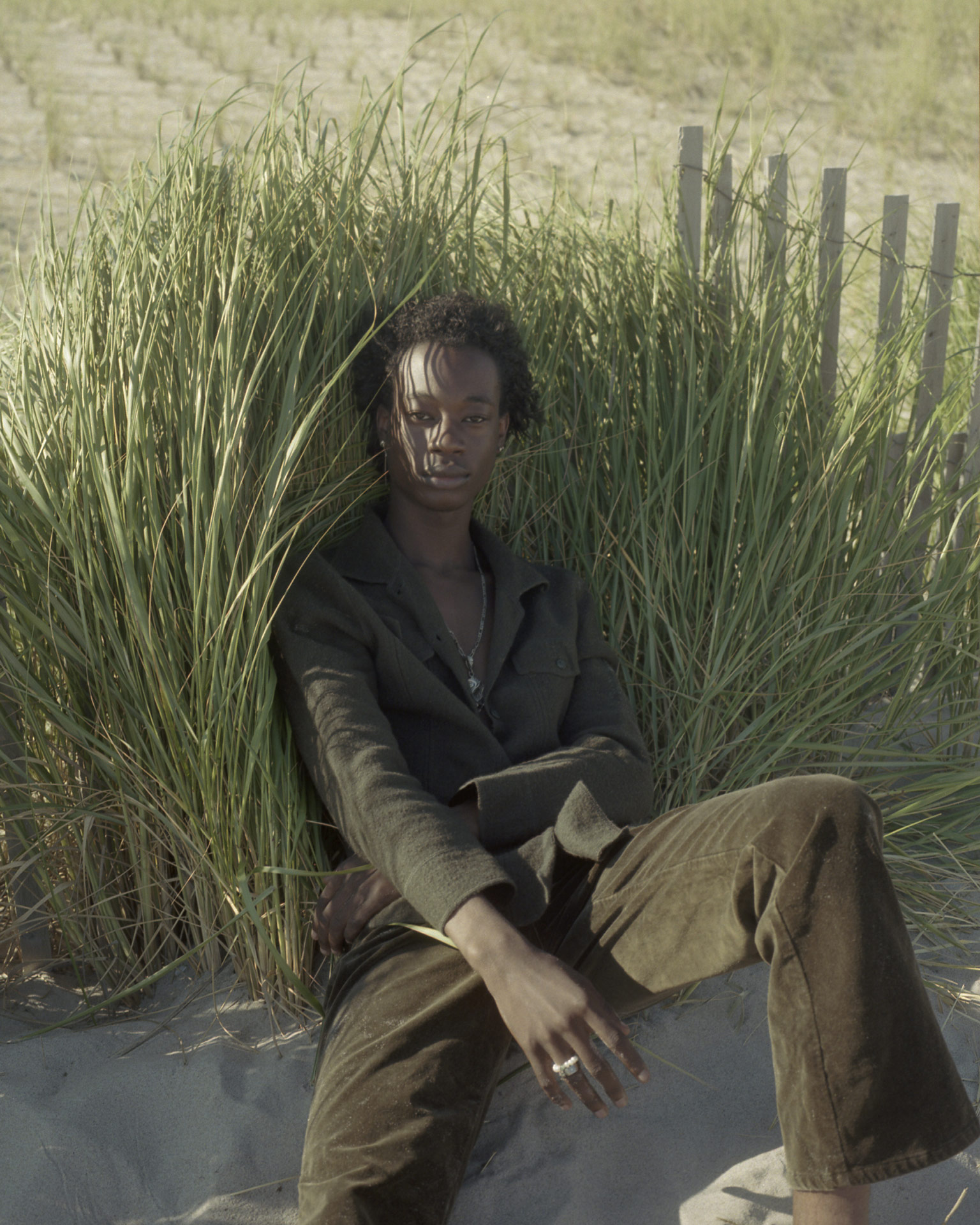 black male sitting in the sand against high grass looking at the camera by Oumayma B. Tanfous for DAZED