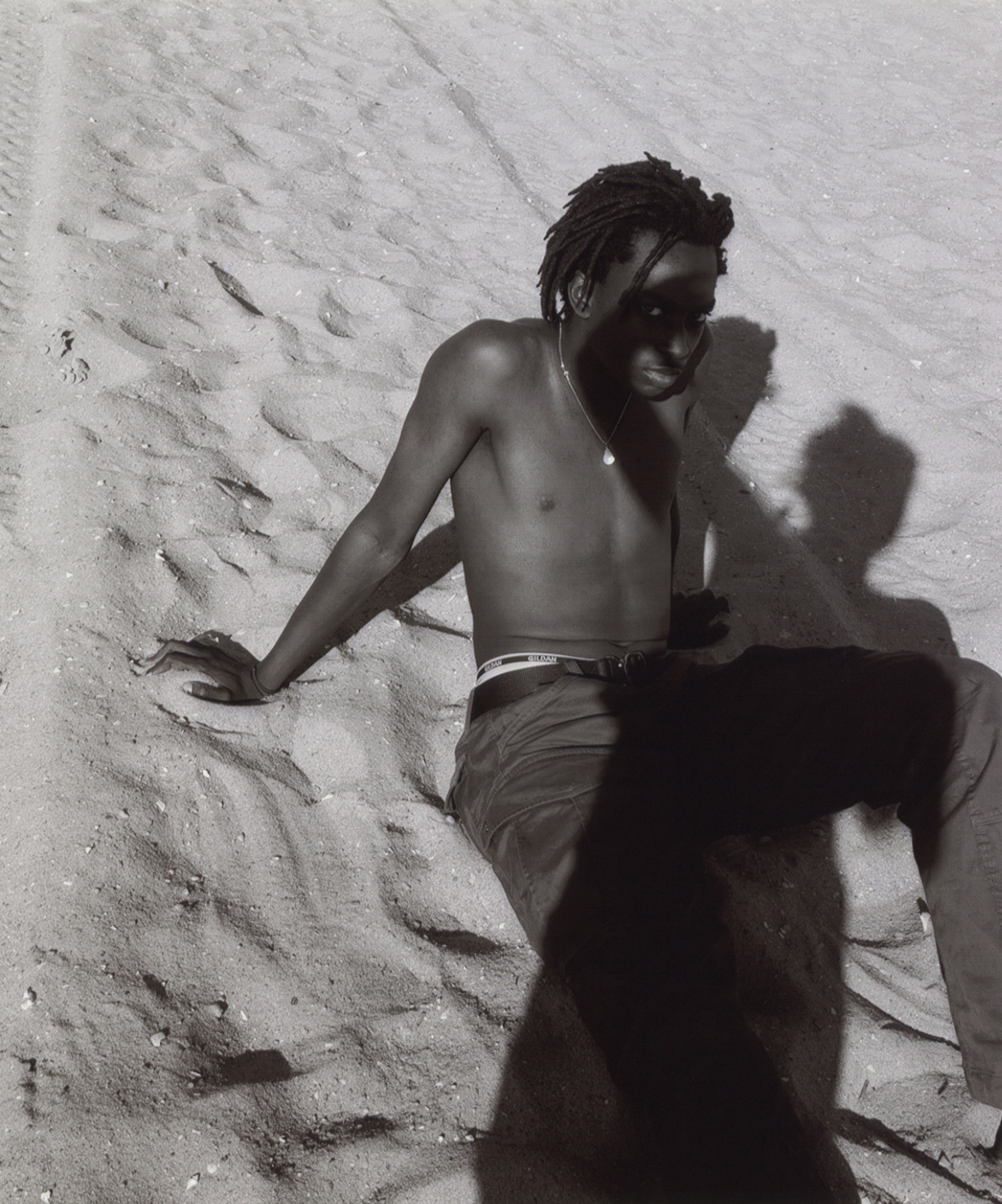 black male sitting in the sand leaning back on his hands shadow of the photographer on him by Oumayma B. Tanfous for DAZED