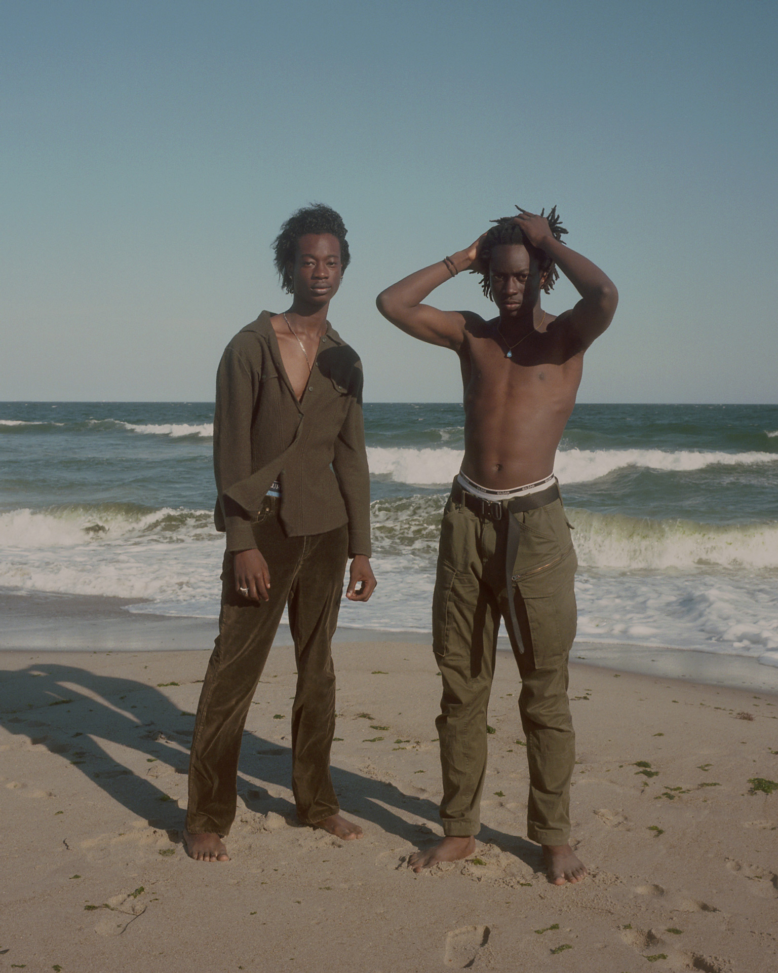 two black male posing on the beach standing up one touching his hair waves crashing behind them by Oumayma B. Tanfous for DAZED