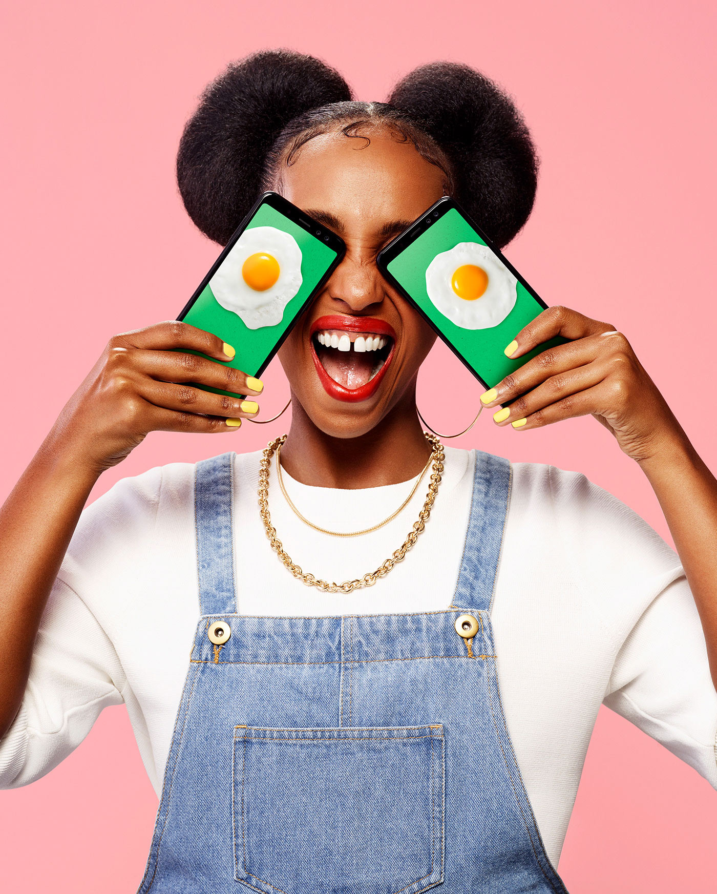 black woman wearing jeans overall and white shirt on pink background with big smile holding two telephones over her eyes displaying pictures of sunny side up eggs by Simon Duhamel for Fizz brand launch with Ogilvy