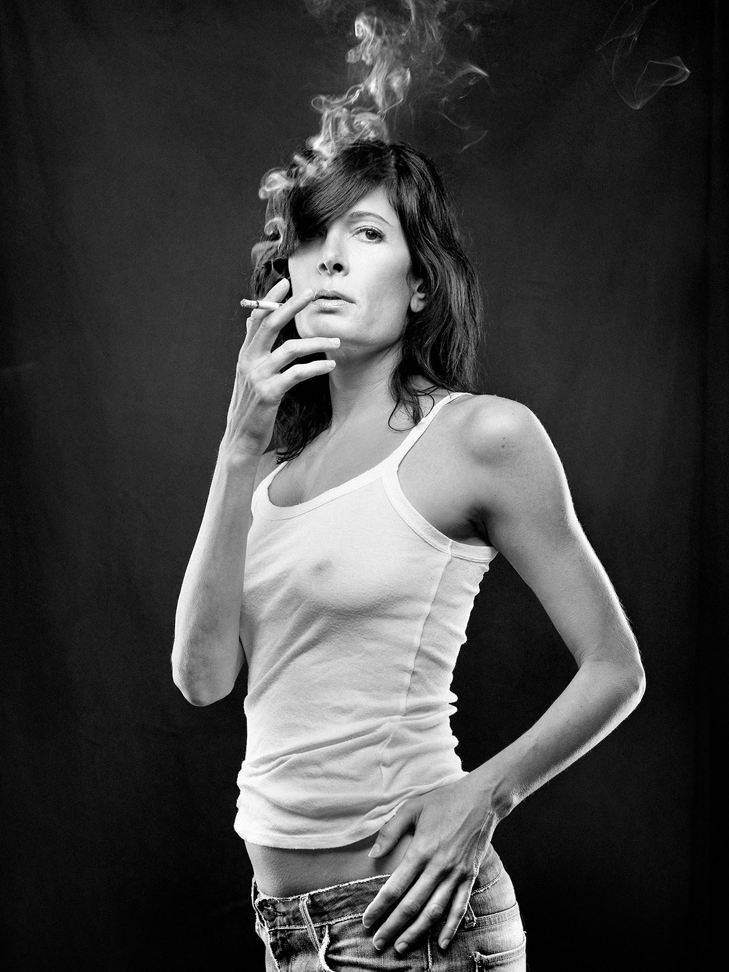 portrait of Anne-Marie Cadieux in black and white wearing white short tank top and low jeans smoking a cigarette on black fabric background by Jocelyn Michel for Voir