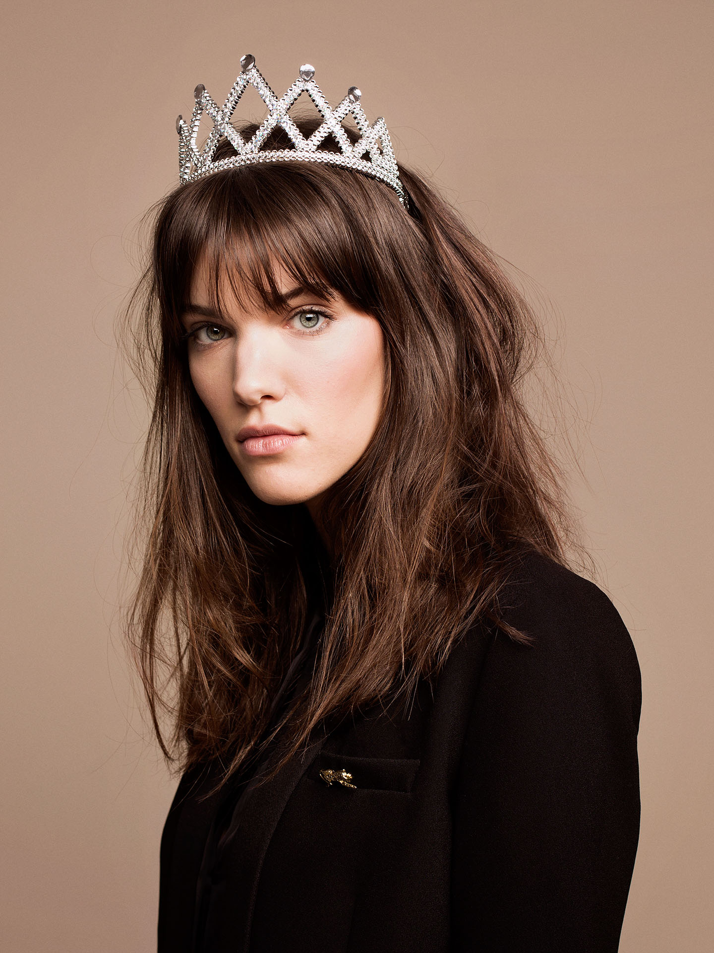 portrait of singer Charlotte Cardin looking at camera dressed in black turtleneck wearing plastic crown on beige background by Jocelyn Michel for Voir