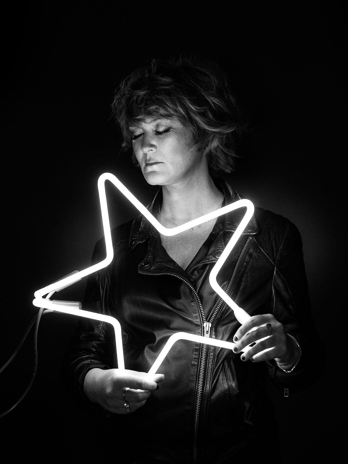 portrait of Mara Tremblay holding lit star-shaped white neon eyes closed in black and white by Jocelyn Michel for Voir